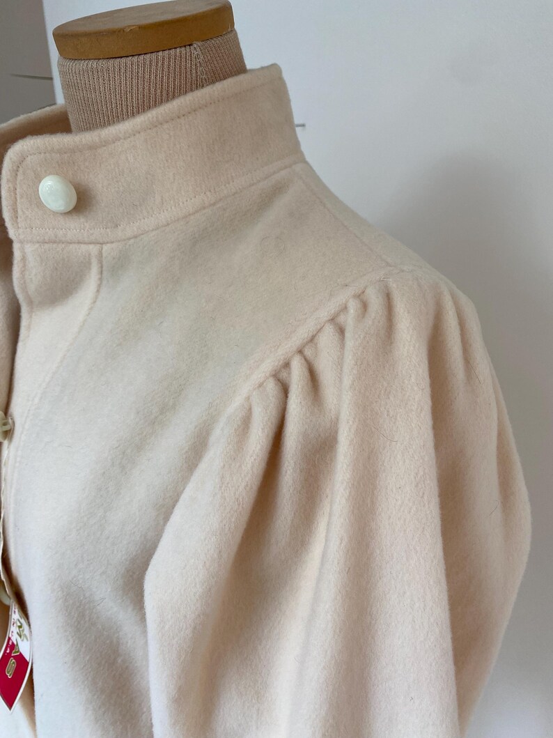 May include: A light peach-colored coat with a high collar and a single large button. The coat features a classic design with gathered sleeves, offering a vintage aesthetic. The fabric appears soft and textured.