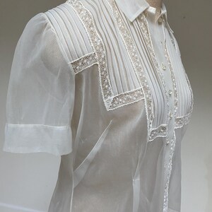 May include: A white sheer blouse with a lace trim and a button-up front. The blouse has a collar and short sleeves.