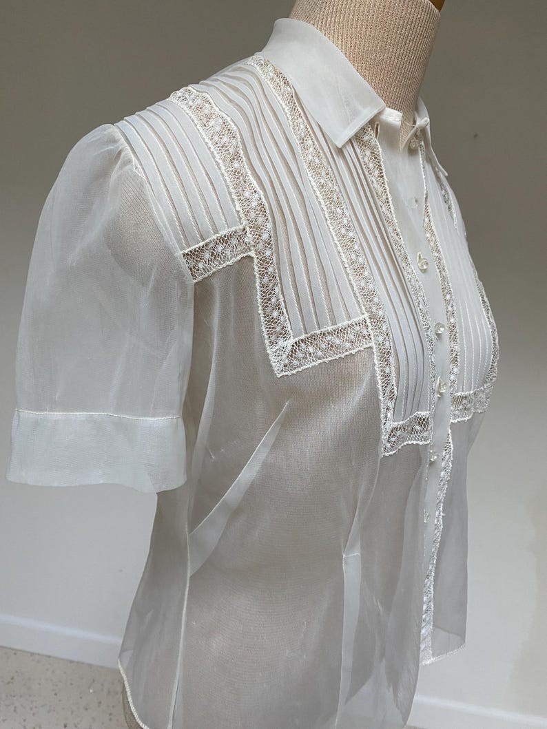 May include: A white sheer blouse with a lace trim and a button-up front. The blouse has a collar and short sleeves.