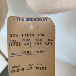 May include: A close-up of a vintage clothing tag from "THE BROADWAY." The tag is beige with black text, displaying numbers and codes such as "025 77110 304," "7201 48," and "4596 9452." The tag is attached to a light-colored garment.