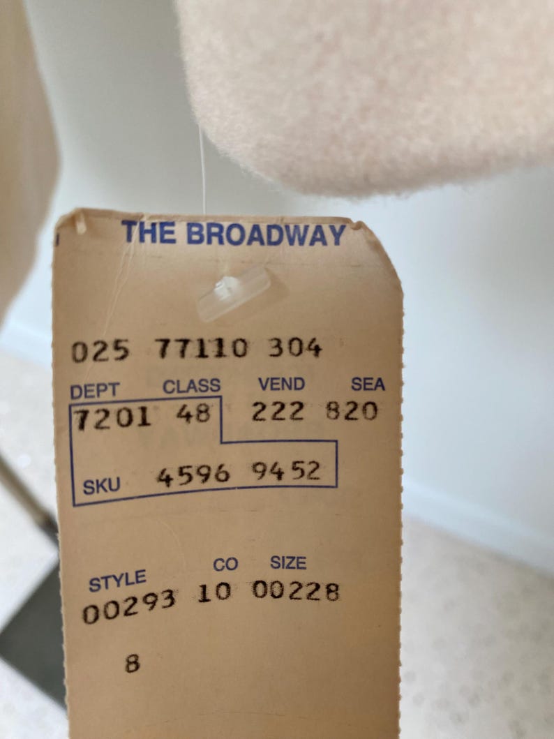 May include: A close-up of a vintage clothing tag from "THE BROADWAY." The tag is beige with black text, displaying numbers and codes such as "025 77110 304," "7201 48," and "4596 9452." The tag is attached to a light-colored garment.