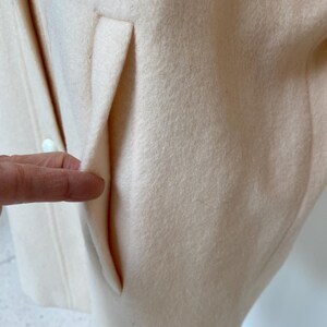 May include: Close-up of a pale peach-colored coat with a pocket detail. The coat appears to be made of a soft, textured fabric. The pocket is a simple, clean design, adding a touch of functionality to the garment.