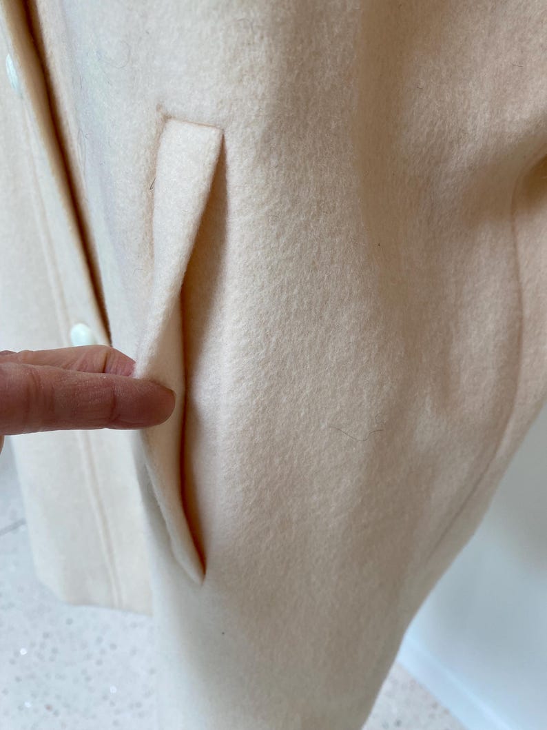 May include: Close-up of a pale peach-colored coat with a pocket detail. The coat appears to be made of a soft, textured fabric. The pocket is a simple, clean design, adding a touch of functionality to the garment.