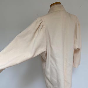May include: A light beige wool coat with a high collar and long sleeves. The coat is displayed on a mannequin, showcasing its tailored silhouette and soft texture. The sleeves have a slight puff at the shoulders.