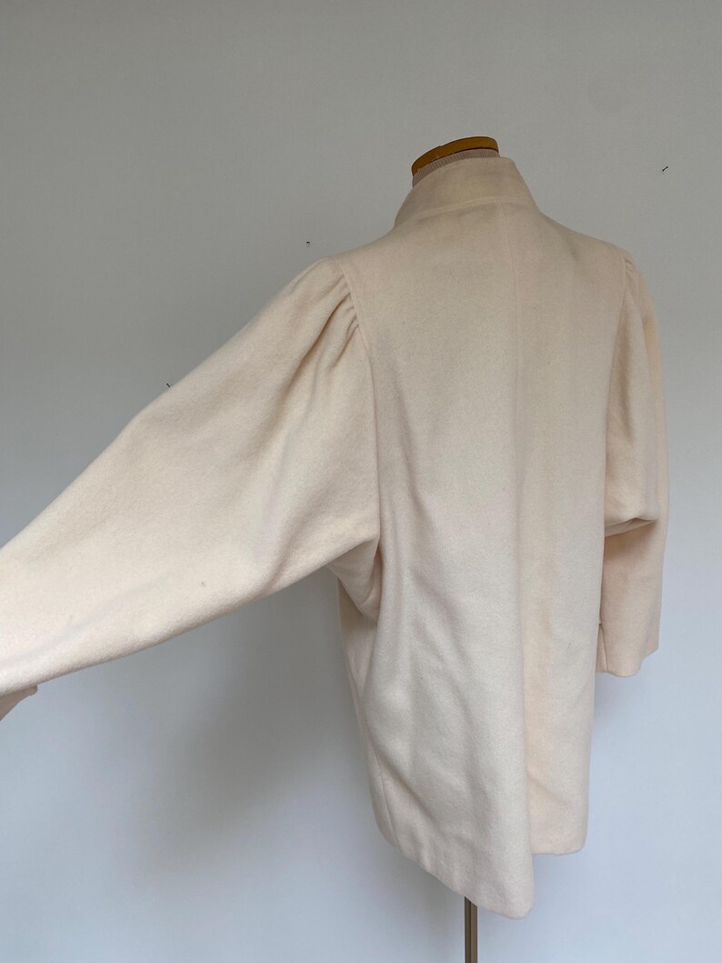 May include: A light beige wool coat with a high collar and long sleeves. The coat is displayed on a mannequin, showcasing its tailored silhouette and soft texture. The sleeves have a slight puff at the shoulders.