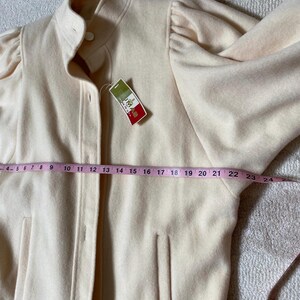 May include: A light beige jacket with a high collar and button closure. The jacket has long sleeves with a gathered detail at the shoulders and two front pockets. A measuring tape shows the jacket is approximately 24 inches long. A tag with text is attached.