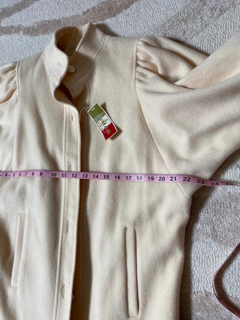 May include: A light beige jacket with a high collar and button closure. The jacket has long sleeves with a gathered detail at the shoulders and two front pockets. A measuring tape shows the jacket is approximately 24 inches long. A tag with text is attached.