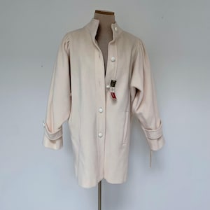 May include: A light pink coat with a high collar and long sleeves. The coat has large white buttons down the front and on the cuffs. A tag hangs from the front. The coat is displayed on a mannequin.