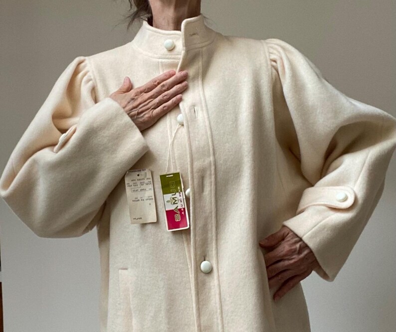 May include: Cream-colored vintage coat with a high collar and button closures. The coat features long sleeves with puffed shoulders and buttoned cuffs. A tag is attached to the coat. The coat is made of a soft, textured fabric.