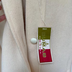 May include: Cream-colored coat with large, round, white buttons. A tag with the text "DUMAS MADE IN U.S.A." is attached. The coat has a classic design and appears to be made of a soft, wool-like material.