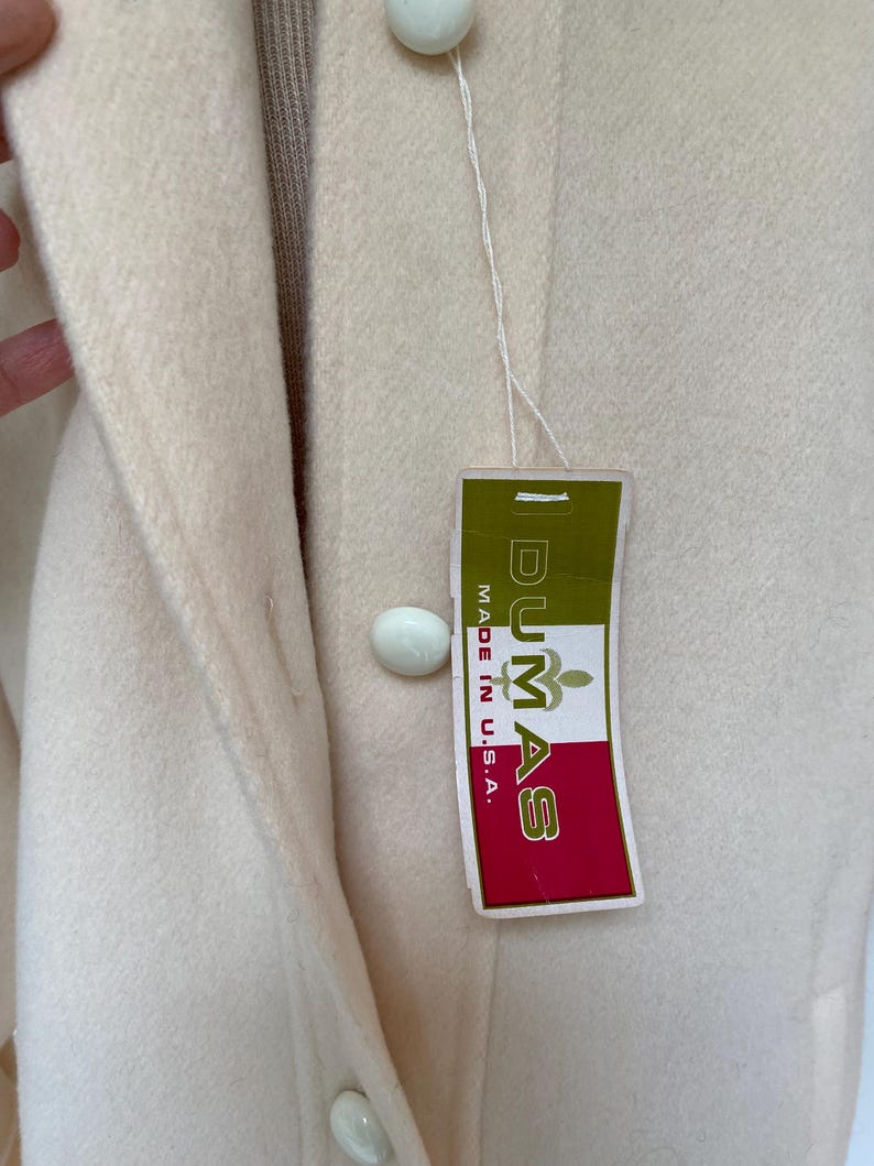 May include: Cream-colored coat with large, round, white buttons. A tag with the text "DUMAS MADE IN U.S.A." is attached. The coat has a classic design and appears to be made of a soft, wool-like material.