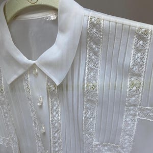 May include: White sheer blouse with a lace trim and a classic collar. The blouse has a button-up front and is pleated throughout.