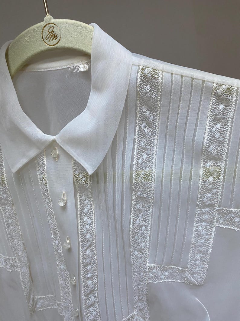 May include: White sheer blouse with a lace trim and a classic collar. The blouse has a button-up front and is pleated throughout.