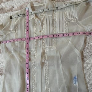 May include: A white sheer blouse with a button-up front and short sleeves. The blouse has a lace trim and is pleated down the front. The blouse is measured with a tape measure, showing it is approximately 19 inches long.