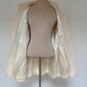 May include: A cream-colored coat displayed on a mannequin. The coat is open, revealing a satin lining in a lighter shade of cream. The coat has a classic design with a collar and a slightly flared shape.