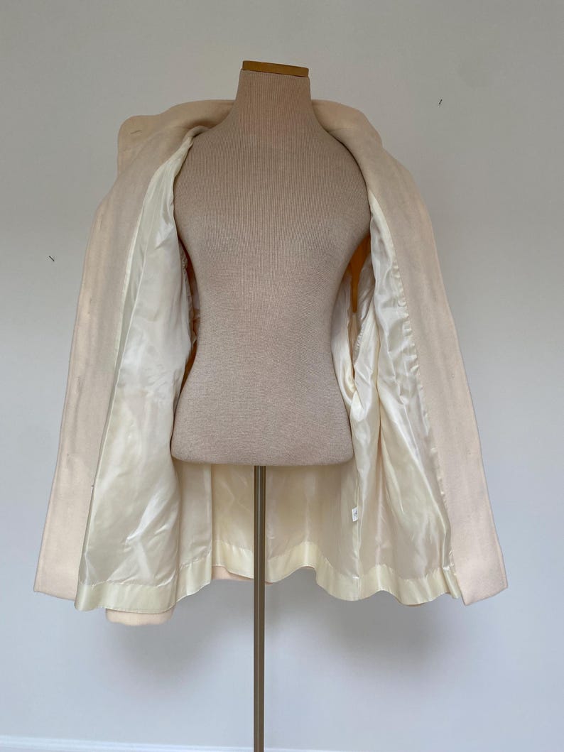 May include: A cream-colored coat displayed on a mannequin. The coat is open, revealing a satin lining in a lighter shade of cream. The coat has a classic design with a collar and a slightly flared shape.