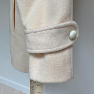 May include: Close-up of a cream-colored coat sleeve with a buttoned cuff. The coat has a classic design with a tailored fit. A price tag hangs from the bottom of the coat.