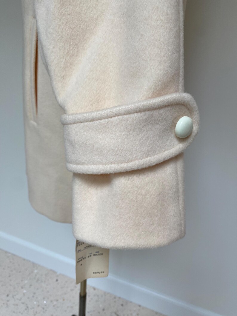 May include: Close-up of a cream-colored coat sleeve with a buttoned cuff. The coat has a classic design with a tailored fit. A price tag hangs from the bottom of the coat.
