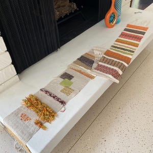 May include: Five woven tapestry samples in various colors and patterns, including brown, orange, green, and white, laid out on a white surface.