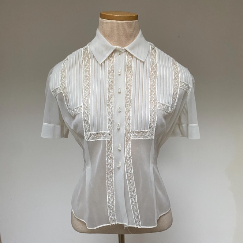 May include: A white sheer blouse with a button-up front and short sleeves. The blouse has lace trim and pleats on the front.