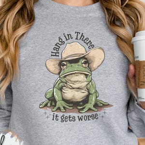 May include: A gray sweatshirt with a green frog wearing a brown cowboy hat. The frog is sitting on a white background with the text "Hang in there it gets worse".