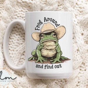 May include: White ceramic mug with a green frog wearing a brown cowboy hat. The frog is sitting on a brown surface and the text "Frog Around and find out" is printed on the mug.