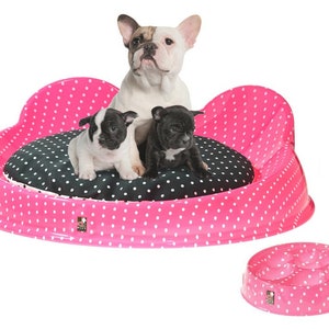 Puede incluir: Una cama para perros de color rosa con lunares blancos y un coj&iacute;n negro con lunares blancos. Tres bulldogs franceses est&aacute;n sentados en la cama. Un cuenco para perros de color rosa con lunares blancos est&aacute; en primer plano.