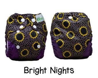 May include: A pair of purple and black cloth diapers with a yellow and black patterned design. The pattern features small circles and larger circles with a yellow ring around them. The text "Kijani" is visible on the diaper. The diapers are labeled "Bright Nights".