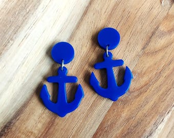 May include: A pair of cobalt blue anchor earrings. Each earring features a circular stud and a dangling anchor shape. The earrings are displayed on a wooden tray with a natural wood grain.