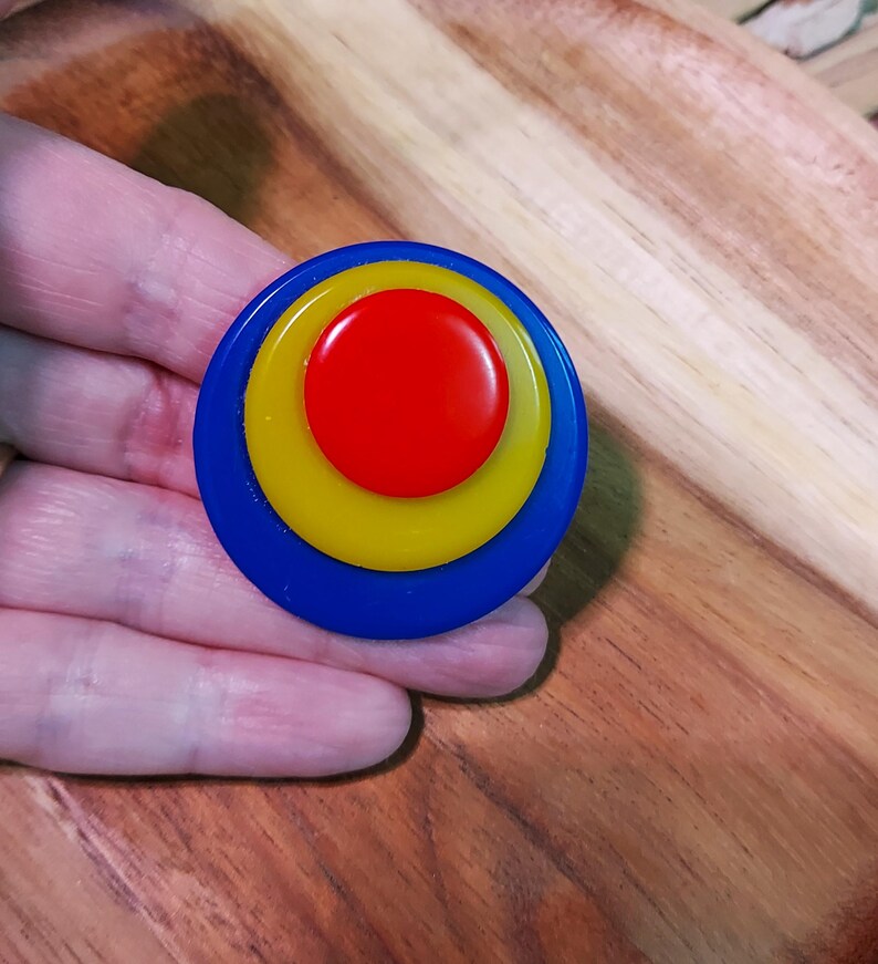 K&ouml;nnte beinhalten: Eine kreisf&ouml;rmige Brosche bestehend aus drei konzentrischen Schichten aus glattem, gl&auml;nzendem Kunststoff. Die Mitte bildet ein roter Kreis, umgeben von einem gelben Ring mittlerer Gr&ouml;&szlig;e und einem gr&ouml;&szlig;eren &auml;u&szlig;eren blauen Ring.