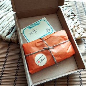 K&ouml;nnte beinhalten: Ein brauner Versandkarton aus Pappe enth&auml;lt ein in orangefarbenes Papier eingewickeltes Paket, das mit Bindfaden verschn&uuml;rt ist. Ein runder Aufkleber zeigt handmade with love mit einem roten Herz. Eine wei&szlig;e Karte mit blauen Punkten tr&auml;gt den Text RosieMays Thank you.