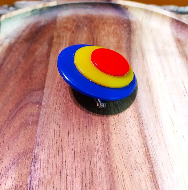 K&ouml;nnte beinhalten: Eine kreisf&ouml;rmige Brosche mit drei konzentrischen Schichten aus glattem, gl&auml;nzendem Kunststoff in Prim&auml;rfarben. Die Mitte bildet ein kleiner roter Kreis, umgeben von einem gelben Ring und einer gr&ouml;&szlig;eren blauen Basis. Ein Metallnadelmechanismus ist auf der Unterseite sichtbar.