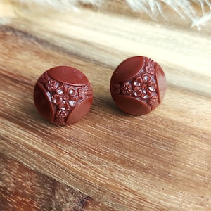 May include: A pair of round, dark reddish-brown buttons featuring a floral pattern. Each button has a raised, detailed design of flowers and leaves. The buttons are displayed on a wooden surface.