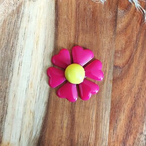 May include: A bright pink flower-shaped brooch with heart-shaped petals and a yellow center. The brooch is set against a wooden background, showcasing its vibrant colors and design. This accessory is perfect for adding a pop of color to any outfit.