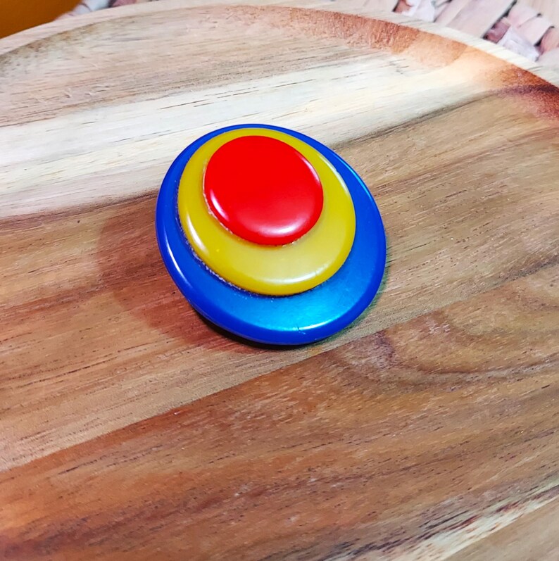K&ouml;nnte beinhalten: Eine kreisf&ouml;rmige Brosche mit drei konzentrischen Schichten aus glattem, gl&auml;nzendem Kunststoff. Die Mitte ist ein roter Kreis, umgeben von einem gelben Ring und einem gr&ouml;&szlig;eren &auml;u&szlig;eren blauen Ring, was ein geschichtetes, mehrfarbiges Design ergibt.