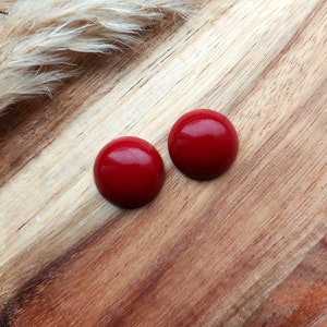 Peut inclure: Deux boucles d'oreilles rondes et brillantes, rouge cerise. Elles sont posées sur un fond en bois, avec une plante douce de couleur neutre dans le coin supérieur gauche. Les boucles d'oreilles ont un design simple et classique.