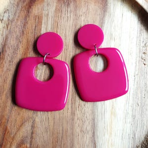 May include: A pair of vibrant pink earrings in a retro style. Each earring has a small, circular stud attached to a larger, square shape with a rounded hole in the middle. The earrings are made of a shiny material.