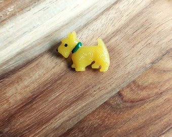 Puede incluir: Una broche de plástico amarilla con forma de perro terrier escocés. El perro lleva un collar verde.