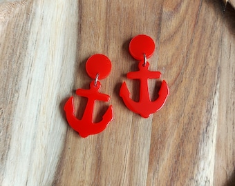 May include: A pair of bright red anchor earrings. Each earring features a circular stud connected to a dangling anchor shape. The earrings are displayed on a wooden surface, creating a nautical theme.