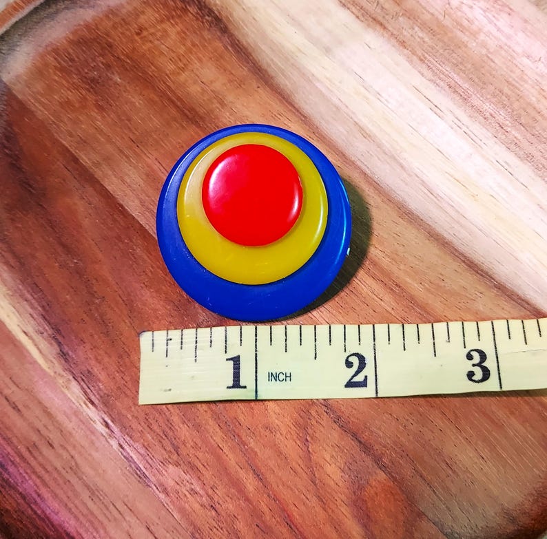 K&ouml;nnte beinhalten: Eine kreisf&ouml;rmige Brosche mit drei konzentrischen Schichten in Rot, Gelb und Blau. Ein gelbes Ma&szlig;band liegt unter der Brosche und zeigt einen Durchmesser von etwa 4,45 Zentimetern f&uuml;r das kreisf&ouml;rmige Objekt an.