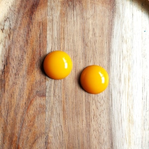 May include: Two glossy, round cabochons in a vibrant yellow hue. They are displayed on a wooden surface, showcasing the natural grain. These could be used for jewellery making or other craft projects.