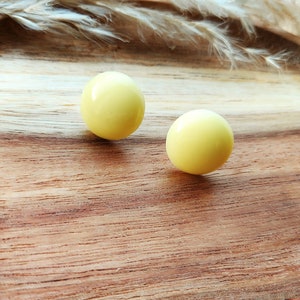 May include: Two pale yellow, spherical earrings sit on a wooden surface. The earrings have a glossy finish and are evenly spaced. The wood grain is visible, and a few dried plants are in the background.