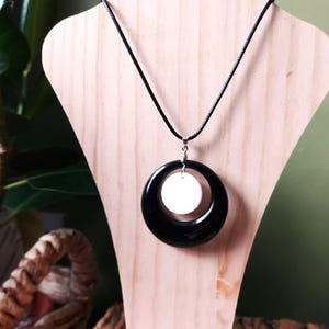 May include: A black and white pendant necklace displayed on a wooden jewelry stand. The pendant features a large black ring with a white circular center, suspended from a black cord.