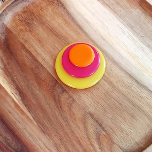 May include: A layered brooch with three circular elements. The largest is yellow, followed by a pink circle, and a small orange circle at the centre. The brooch is set against a wooden background.