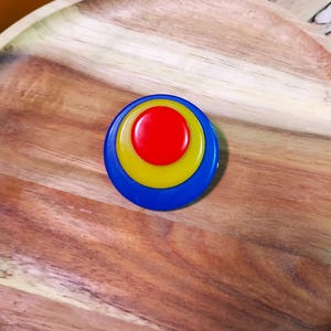 K&ouml;nnte beinhalten: Eine kreisf&ouml;rmige Brosche mit drei konzentrischen Farbringen. Die Mitte ist ein roter Kreis, umgeben von einem gelben Ring und einem &auml;u&szlig;eren blauen Ring. Die Oberfl&auml;che der Brosche ist glatt und gl&auml;nzend, was ein geschichtetes Erscheinungsbild erzeugt.