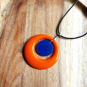 May include: A striking orange pendant necklace with a circular design, featuring a smaller blue circle within the larger orange one. The pendant hangs from a black cord, set against a wooden background.