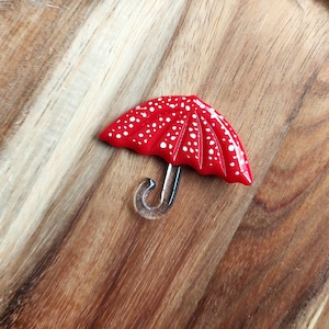 May include: A red umbrella-shaped brooch with white polka dots. The handle is clear and curved. The brooch is set against a wooden background.