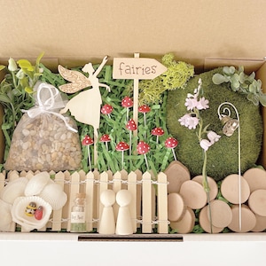 May include: A fairy garden kit presented in a cardboard box. The kit contains a wooden fairy, a sign reading "fairies", miniature red and white toadstools, a white picket fence, and a bag of pebbles. Small plants and a moss-covered sphere are also included.