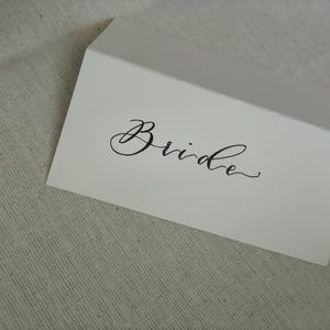 May include: A white card, folded at an angle, with the word "Bride" written in black cursive script. The card sits on a textured, light-coloured fabric.