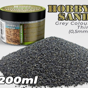 Op de afbeelding: Een pot van 200 ml grijze hobbyzand, met het label "Hobby Sand Grey Colour Thin (0,5mm)". Het zand is fijn en dun, ideaal voor modelbouw en terreinbouw.